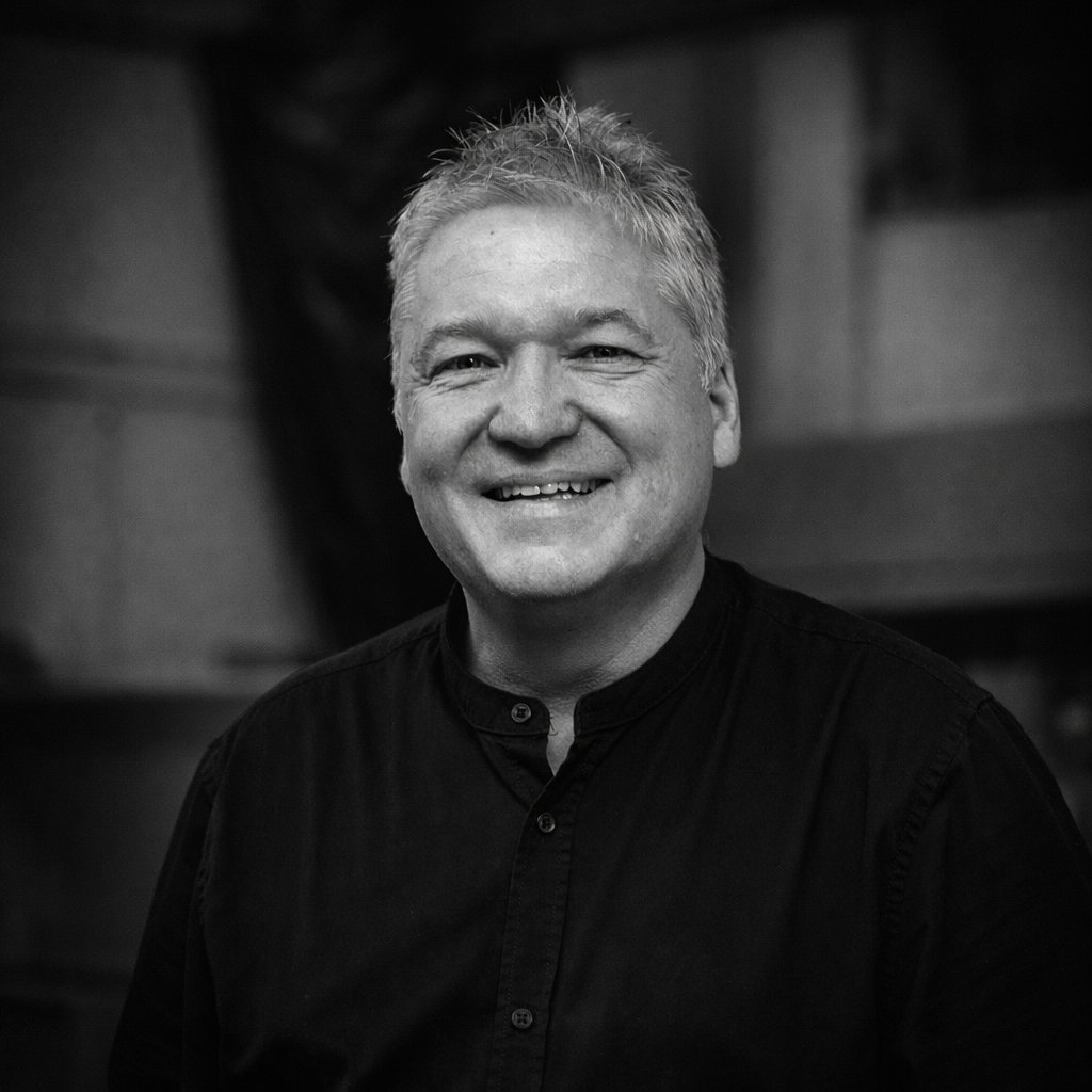 Porträt von Mario Esch in schwarzem Stehkragenhemd, lachend vor dem unscharfen Hintergrund seiner Schreinerwerkstatt — Schwarz-Weiß-Aufnahme